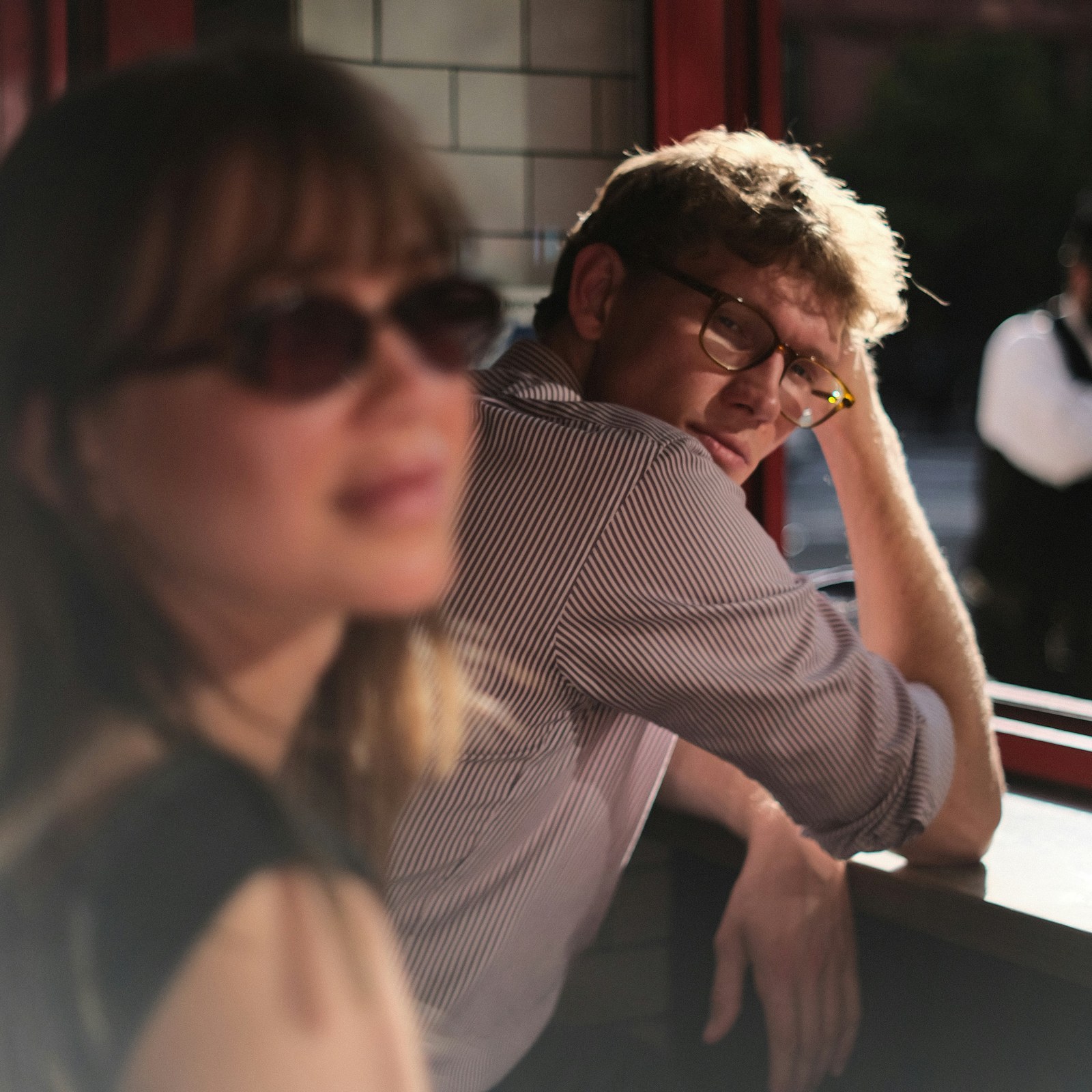 A man sits at a bar with his head resting on his hand, looking past a woman who is out of focus in the foreground.